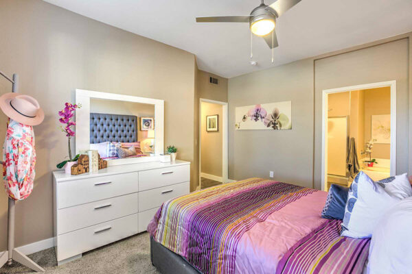 Cozy bedroom in apartment in Summerlin with carpet, plush bed with dresser with mirror, coat rack, and door to bathroom.