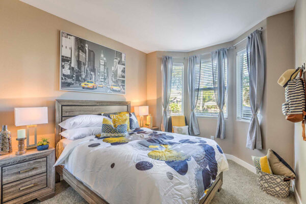 Large bedroom in Summerlin apartment with carpet, wood bed with headboard, bedside drawers with lamps, artwork, and large windows.