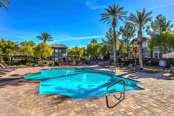 Pool area with stone paver patio, lounge chairs, lush landscaping, and tall palm trees.