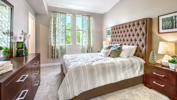 Bedroom with carpet, plush bed with headboard, dressers with lamps and decor, and large windows.