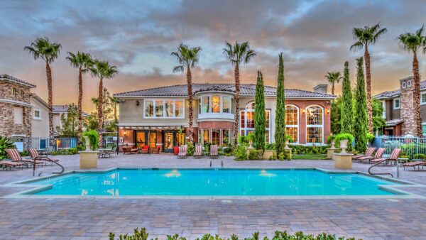 Pool area with stone paver patio, lounge chairs, lush landscaping, and tall palm trees.