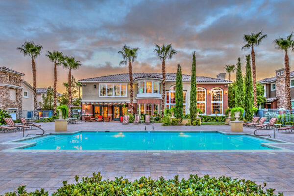 Pool area with stone paver patio, lounge chairs, lush landscaping, and tall palm trees.