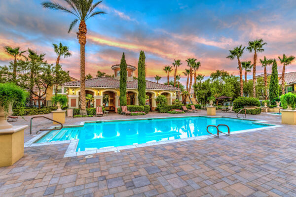 Pool area with stone paver patio, lounge chairs, lush landscaping, and tall palm trees.