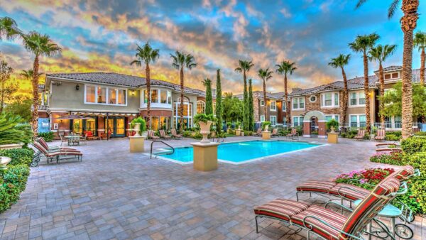 Pool area with stone paver patio, lounge chairs, lush landscaping, and tall palm trees.