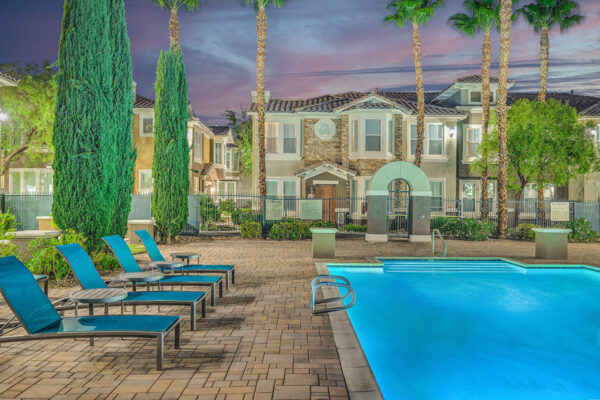 Pool area with stone paver patio, lounge chairs, lush landscaping, and tall palm trees.