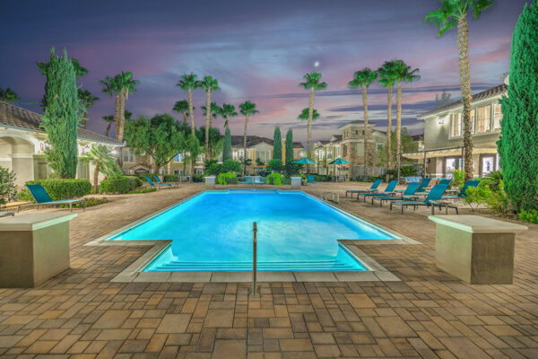 Pool area with stone paver patio, lounge chairs, lush landscaping, and tall palm trees.