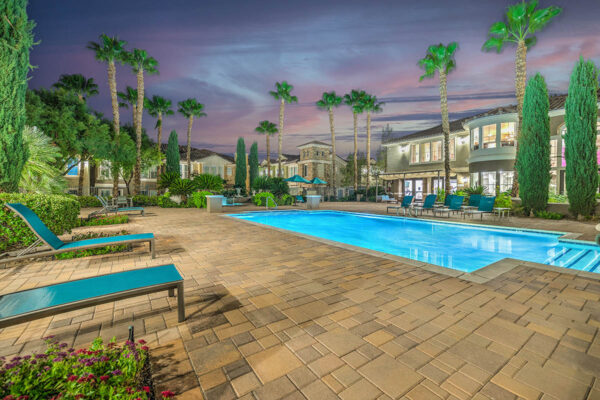 Pool area with stone paver patio, lounge chairs, lush landscaping, and tall palm trees.