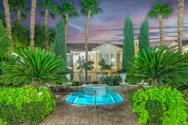 Pool area with stone paver patio, lounge chairs, lush landscaping, and tall palm trees.