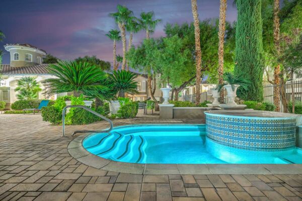 Pool area with stone paver patio, tiled spa, lush landscaping, and tall palms.