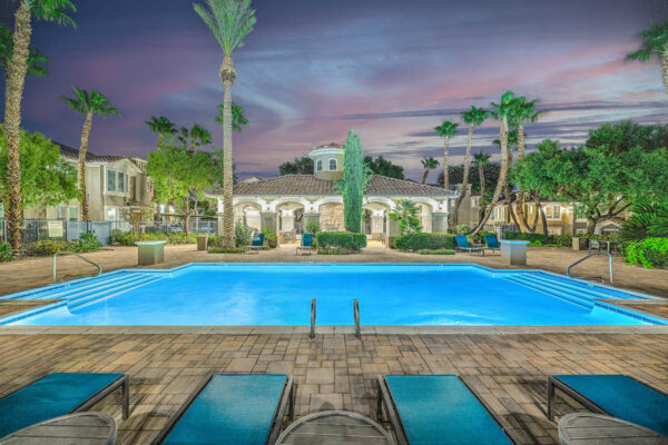 Pool area with stone paver patio, lounge chairs, lush landscaping, and tall palm trees.