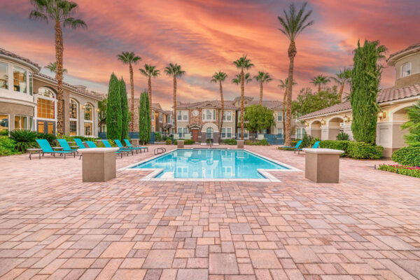 Pool area with stone paver patio, lounge chairs, lush landscaping, and tall palm trees.