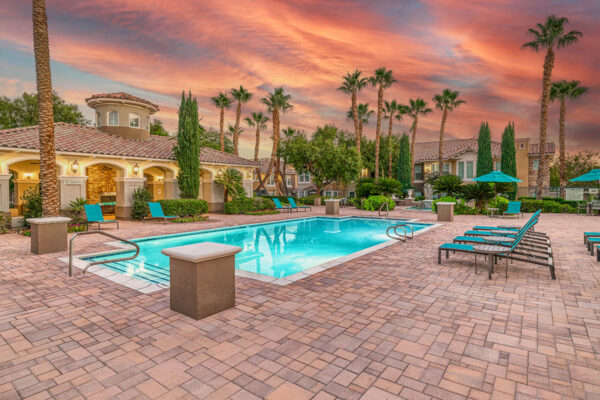 Pool area with stone paver patio, lounge chairs, lush landscaping, and tall palm trees.
