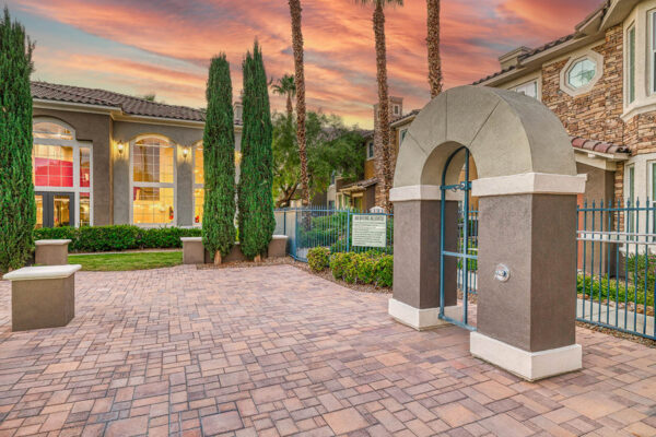 Outdoor area with stone paver patio, archway, lush landscaping, and tall palm trees.