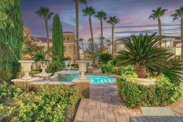 Pool area with stone paver patio, lounge chairs, lush landscaping, and tall palm trees.