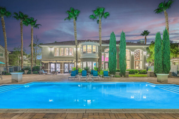 Pool area with stone paver patio, lounge chairs, lush landscaping, and tall palm trees.