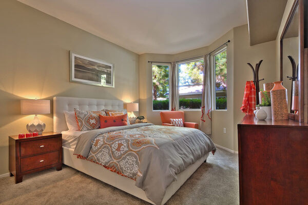 Bedroom with gray and orange patterned bedding, dark wood dresser, and large bay windows.