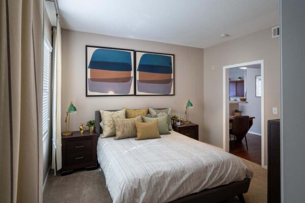 Arzano apartment bedroom with carpet, plush bed with pillows, bedside tables with lamps, and framed art.